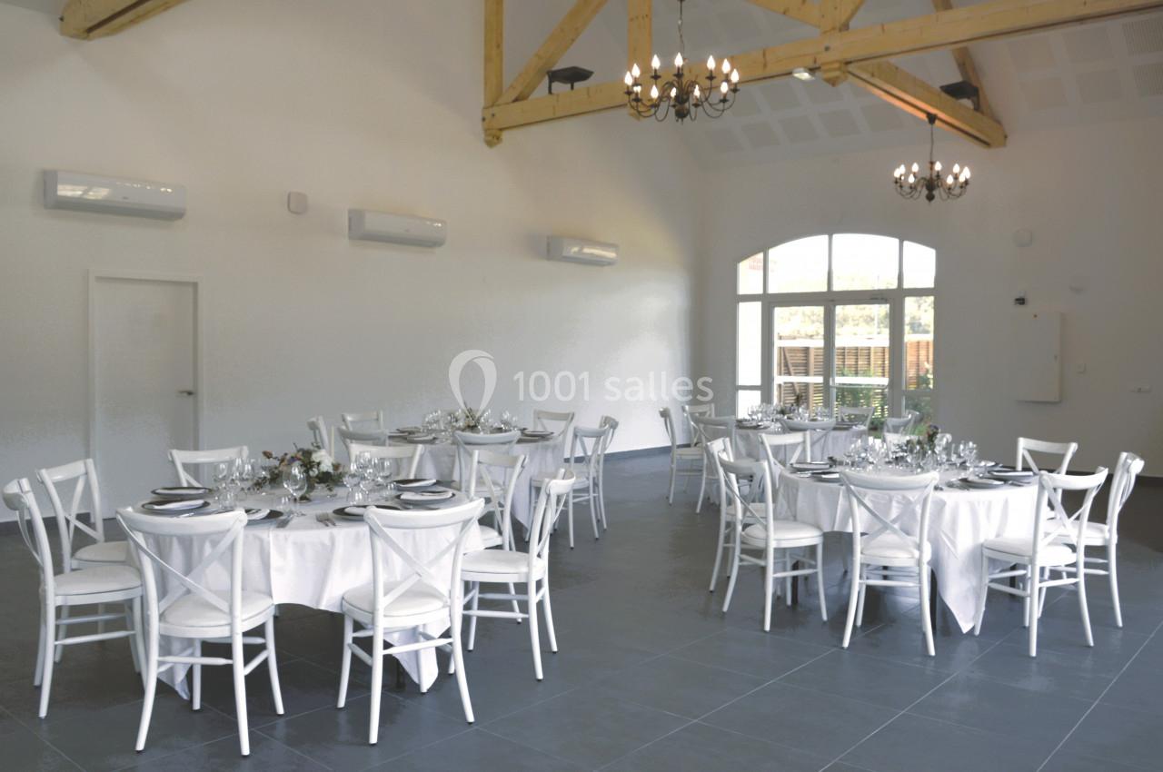 Salle de réception lumineuse avec tables rondes dressées, chaises blanches et poutres apparentes au plafond.