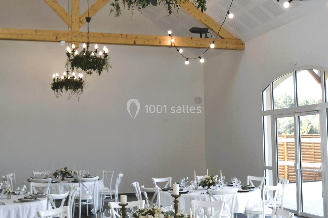 Salle de réception lumineuse avec tables rondes dressées, chaises blanches et poutres apparentes en bois.