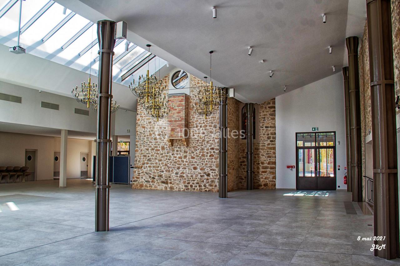 Salle spacieuse avec murs en pierre, grandes baies vitrées, lustres suspendus et sol en carrelage gris.