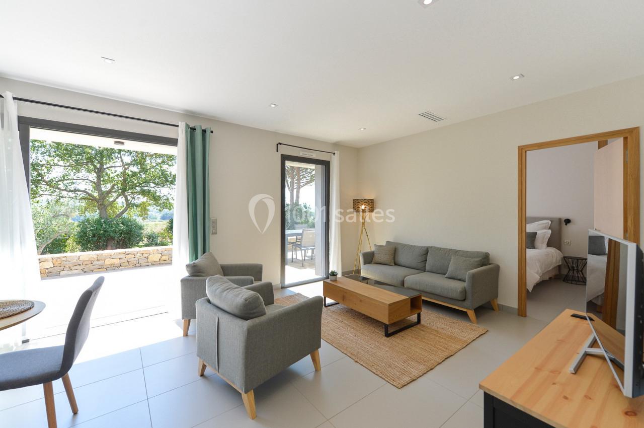 Salon lumineux avec canapé, fauteuils, table basse, ouvert sur une terrasse et une chambre visible en arrière-plan.