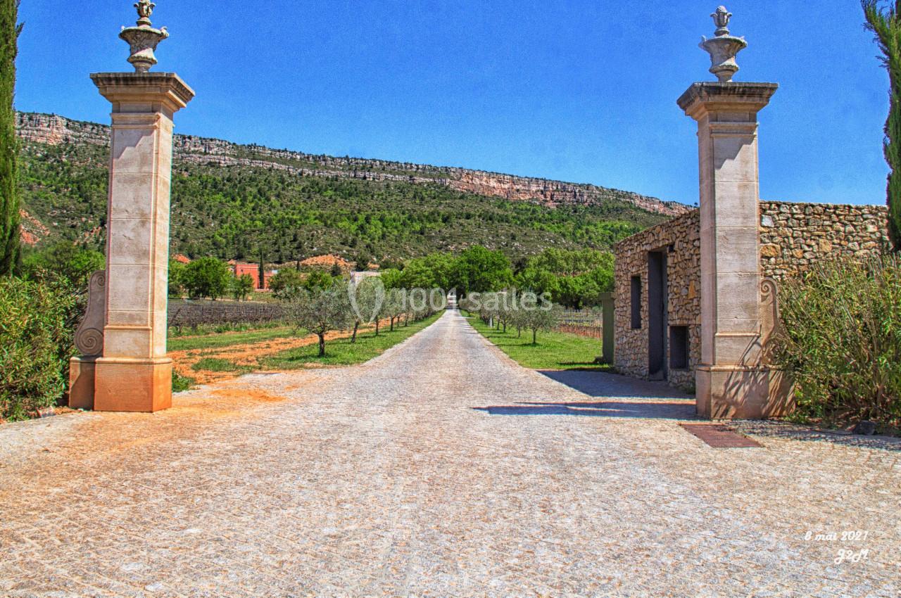 Allée bordée d'arbres et de champs, encadrée par deux piliers en pierre, menant vers un paysage vallonné.