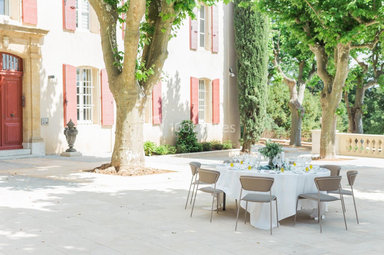Table dressée en extérieur sous des arbres, devant une bâtisse aux volets rouges, par une journée ensoleillée.