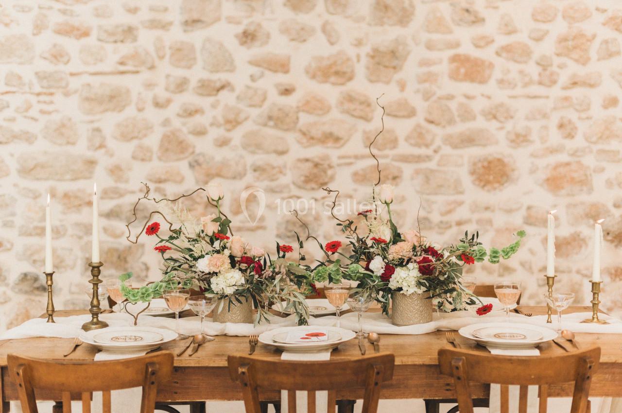 Table en bois décorée de fleurs colorées, bougies et vaisselle élégante, devant un mur en pierre apparente.
