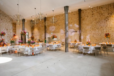 Salle de réception lumineuse avec murs en pierre, tables décorées de fleurs colorées et chandeliers suspendus.