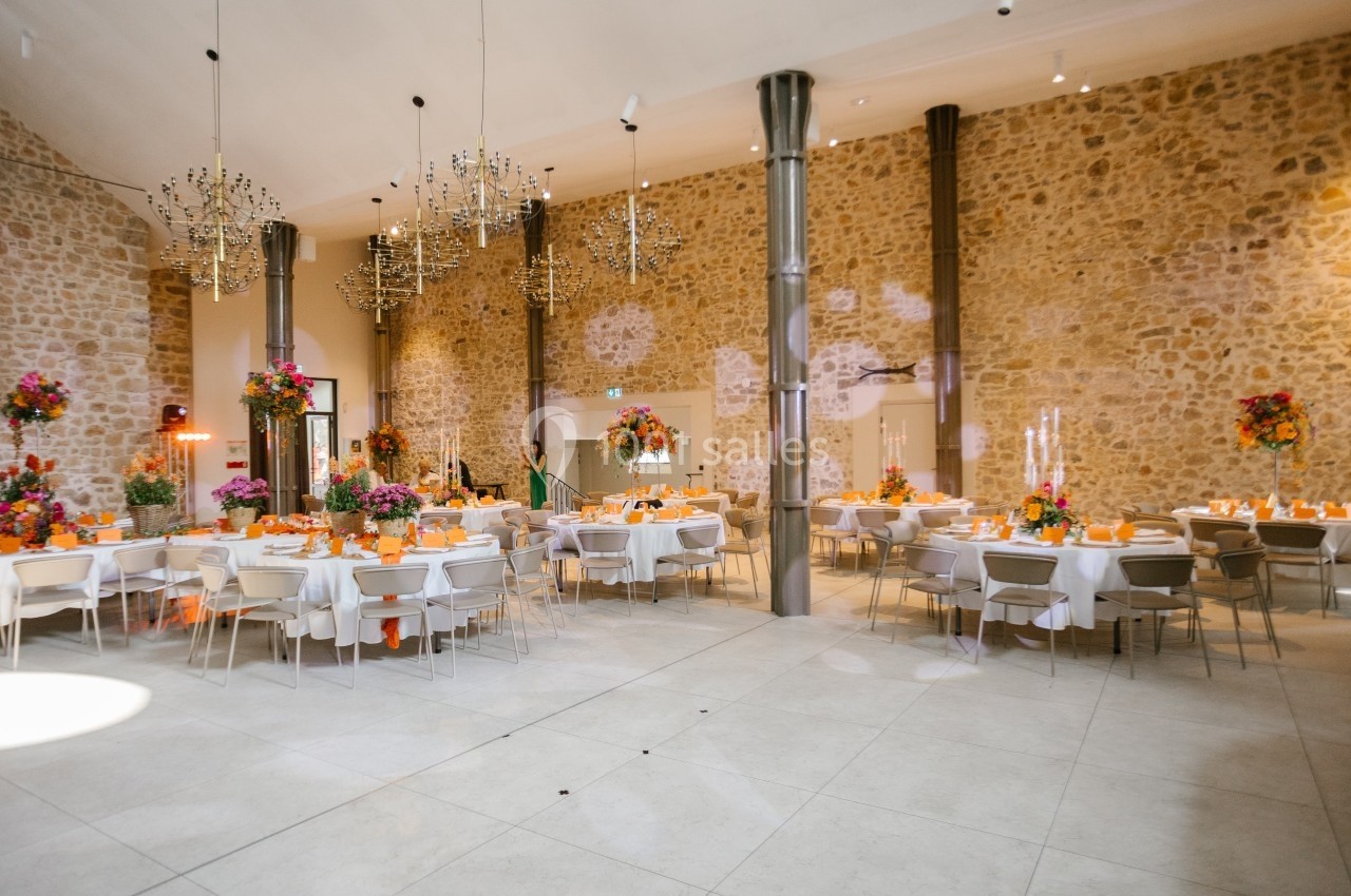 Salle de réception lumineuse avec murs en pierre, tables décorées de fleurs colorées et chandeliers suspendus.