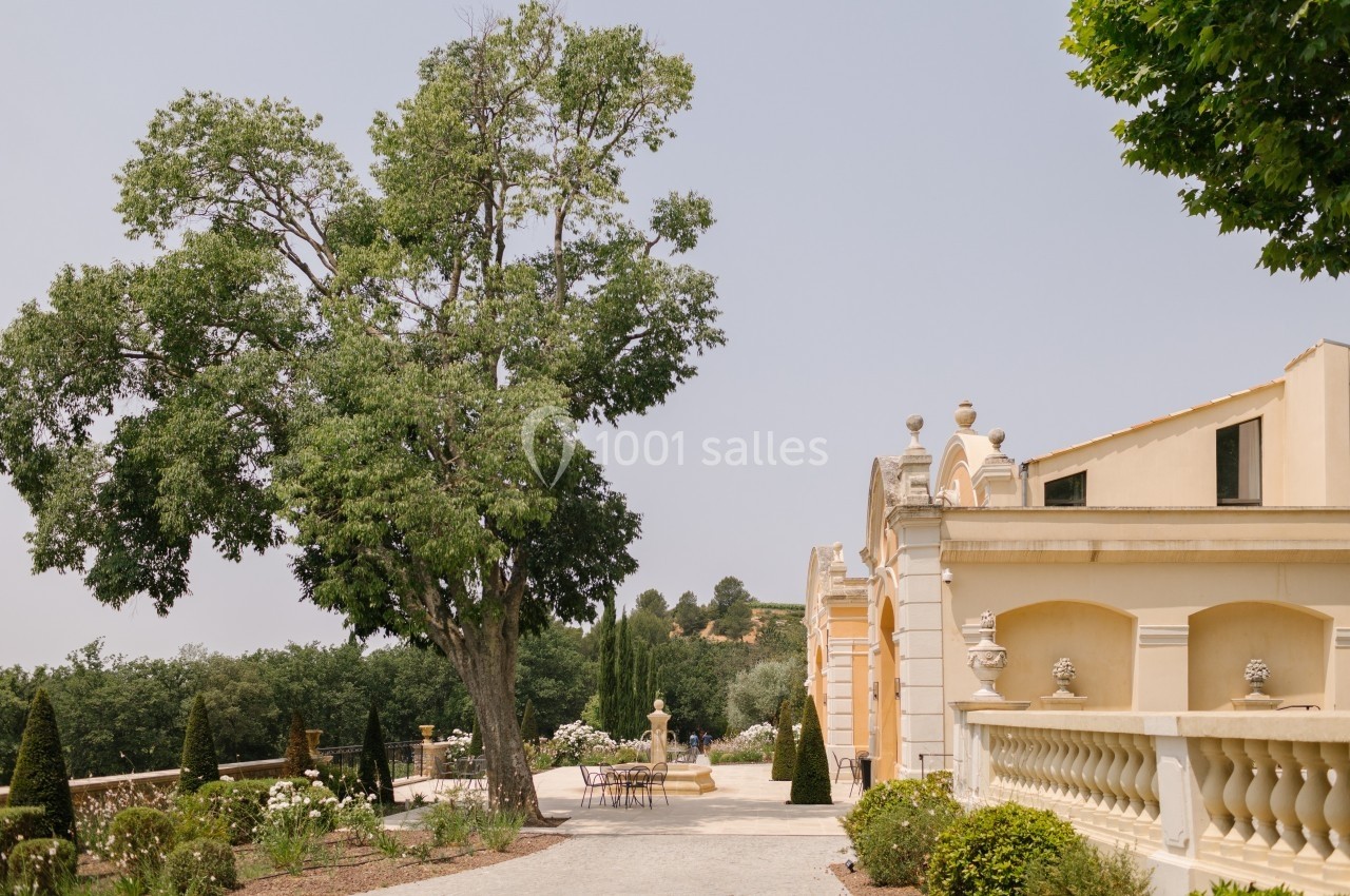 Allée bordée de verdure menant à une bâtisse beige avec balustrades, entourée d'arbres et d'arbustes.