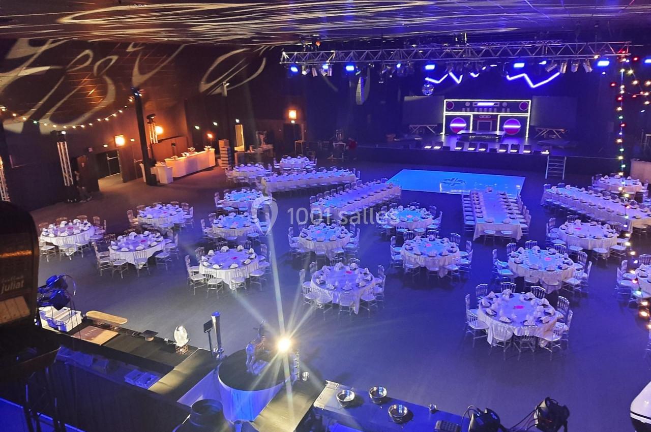 Salle de réception avec tables rondes dressées, éclairages colorés et scène équipée pour un événement.