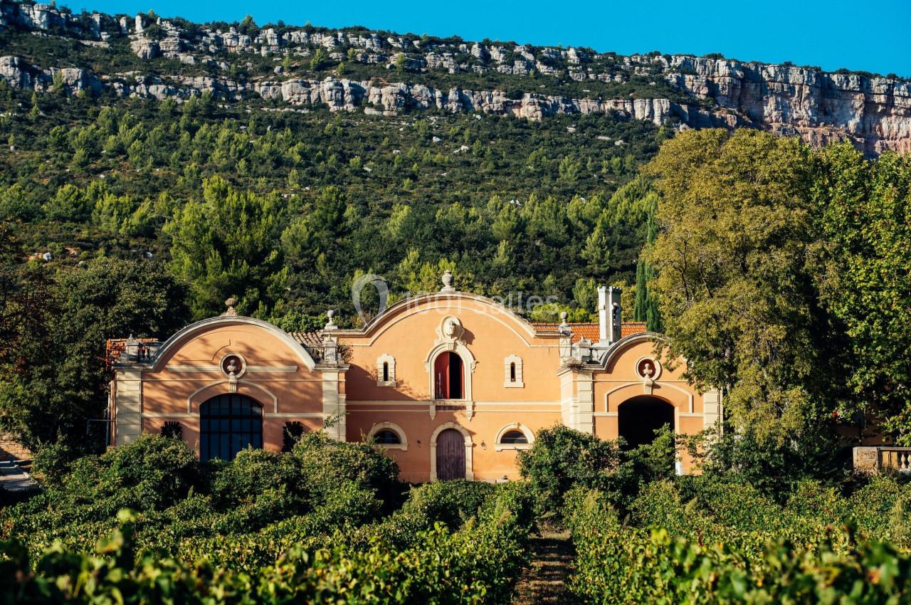 Bâtiment aux façades orangées entouré de vignes, avec une colline boisée en arrière-plan sous un ciel bleu.