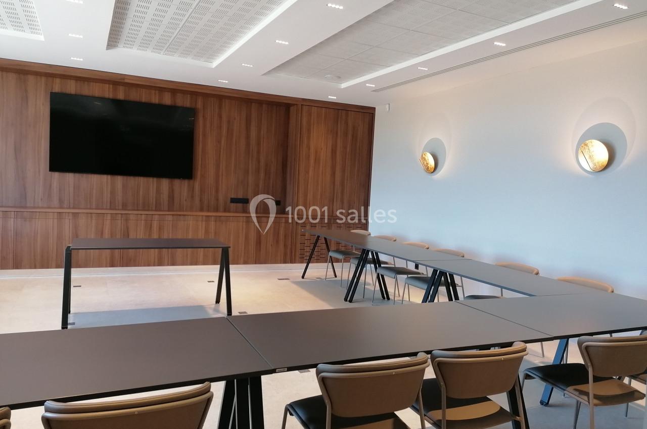 Salle de réunion moderne avec tables en U, chaises rembourrées, mur en bois, écran mural et éclairage discret.