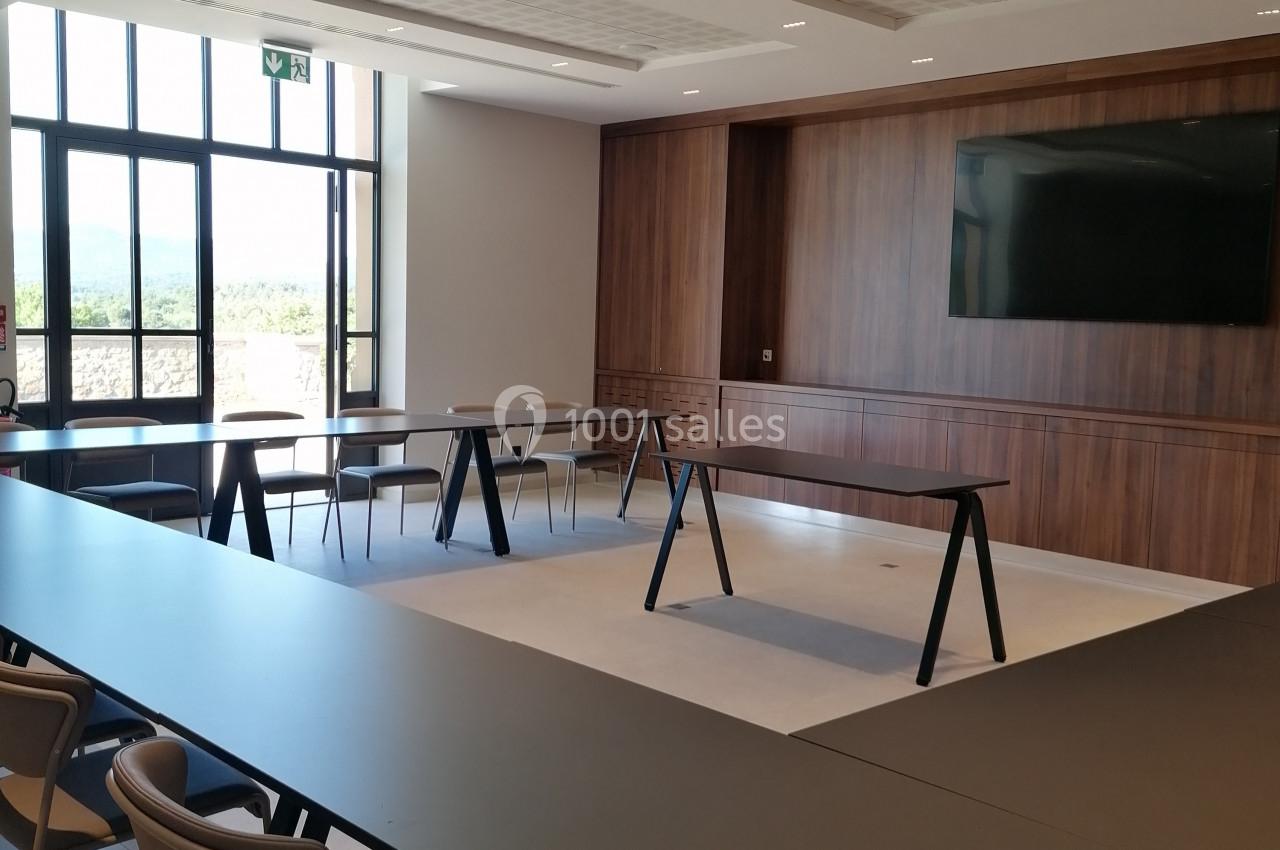 Salle de réunion moderne avec tables en U, chaises, écran mural et grande fenêtre laissant entrer la lumière naturelle.