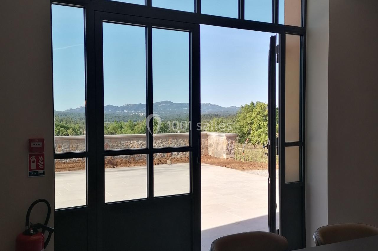 Vue sur un paysage extérieur depuis une salle avec une grande baie vitrée ouverte, table et chaises au premier plan.