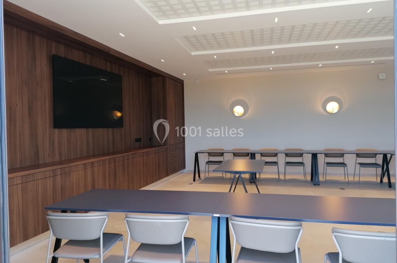 Salle de réunion moderne avec tables, chaises alignées et écran mural, éclairée par des appliques murales.