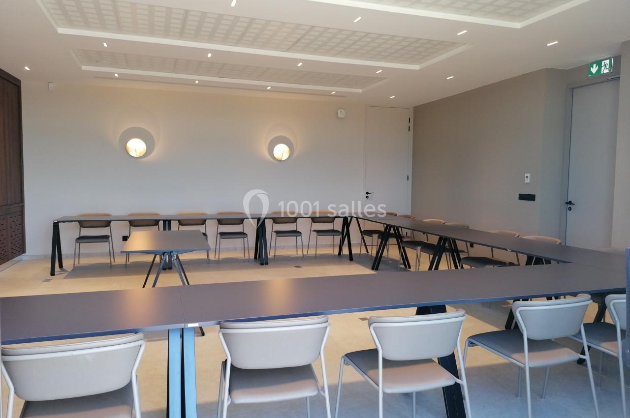 Salle de réunion lumineuse avec tables disposées en U et chaises beige clair, murs neutres et éclairage mural.