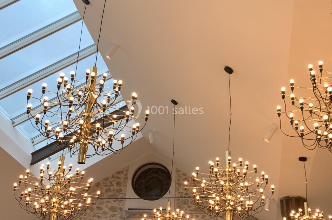 Plafond avec lustres dorés suspendus, mur en pierre apparente et verrière laissant entrer la lumière naturelle.