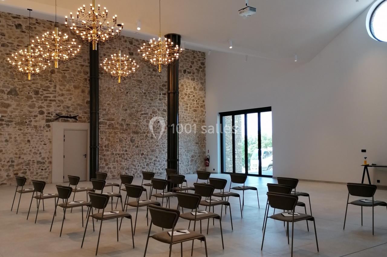 Salle lumineuse avec murs en pierre, chaises disposées en rangées, grandes baies vitrées et lustres modernes suspendus.