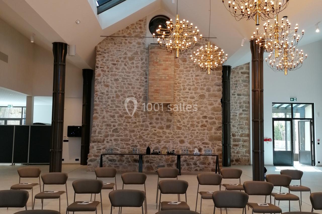 Salle lumineuse avec murs en pierre, chaises disposées en rangées et lustres suspendus sous un plafond vitré.