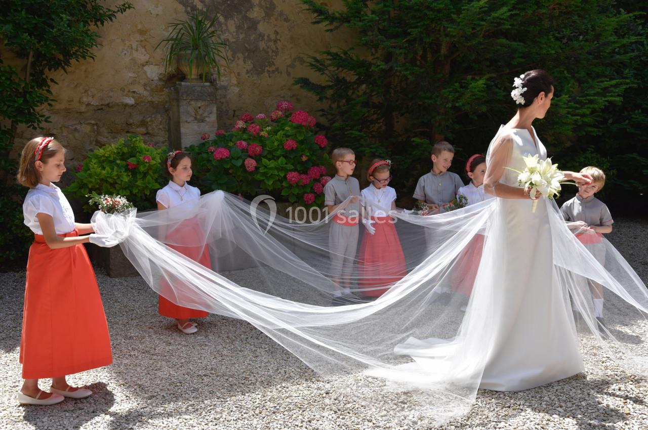 Une mariée en robe blanche avec un long voile tenu par des enfants d'honneur dans un jardin fleuri.