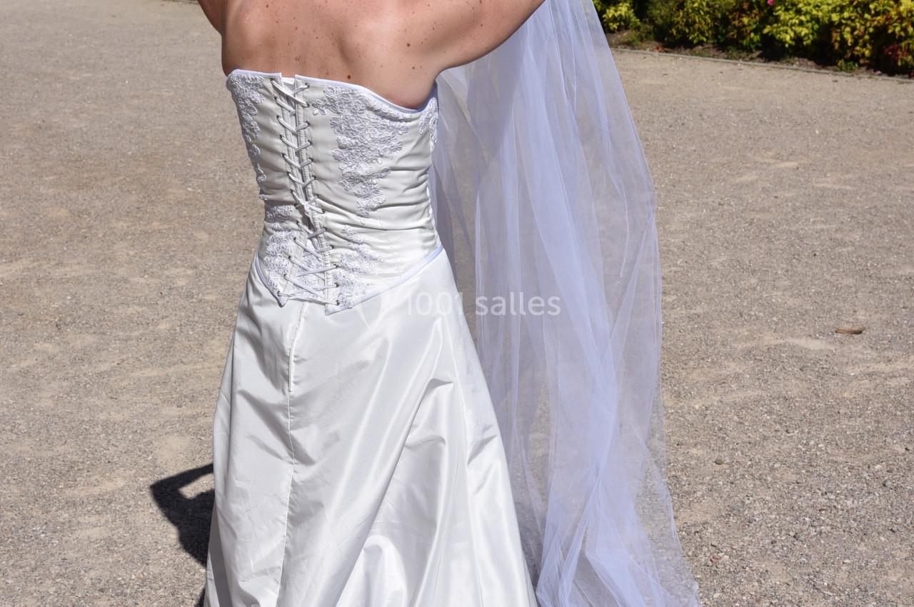 Une femme en robe de mariée blanche ajuste un long voile dans un jardin fleuri sous un ciel ensoleillé.