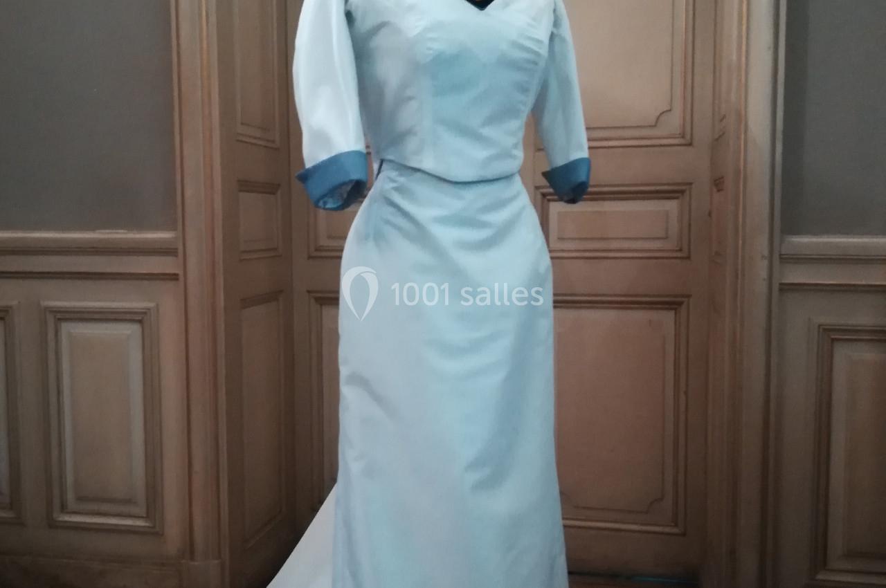 Robe longue blanche avec manches trois-quarts et détails bleus, présentée sur un mannequin dans une pièce élégante.