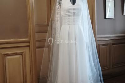 Mannequin vêtu d'une robe en dentelle blanche avec un voile noir couvrant la tête, devant une porte en bois clair.