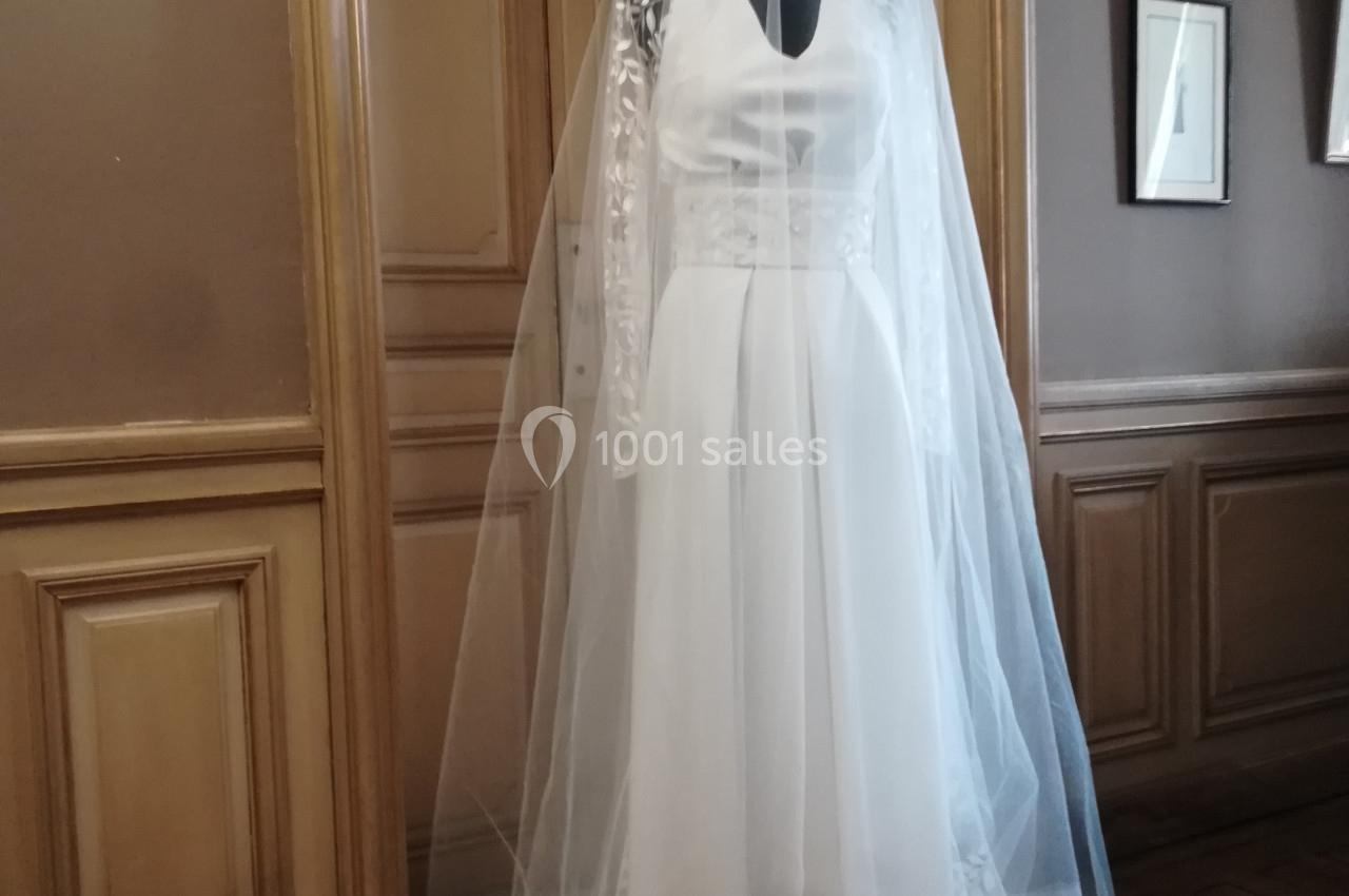 Mannequin portant une robe de mariée blanche avec un long voile, placé devant une porte en bois dans une pièce élégante.