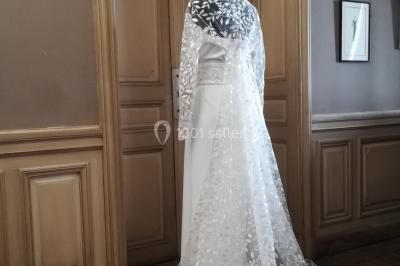 Mannequin vêtu d'une robe en dentelle blanche avec un voile noir couvrant la tête, devant une porte en bois clair.