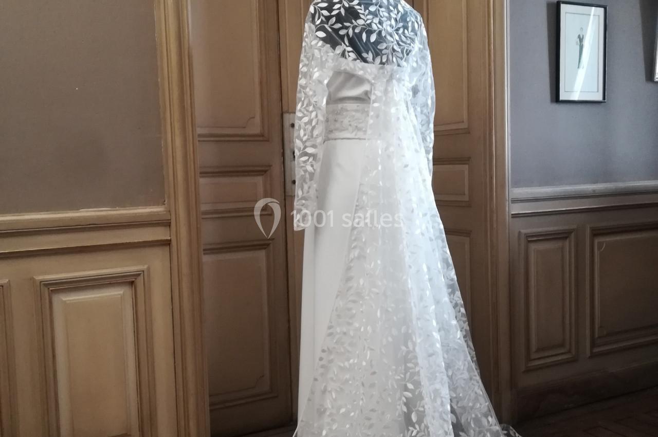 Mannequin vêtu d'une robe de mariée blanche avec une longue cape en dentelle florale, dans une pièce élégante.