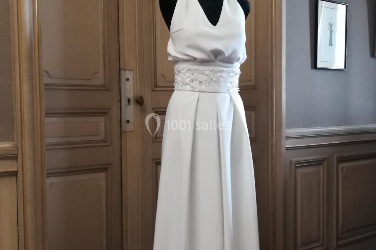 Mannequin portant une robe de mariée blanche sans manches avec ceinture ornée, dans une pièce au décor classique.