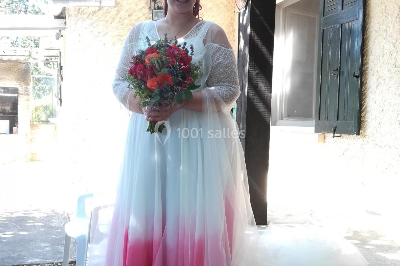 Femme en robe de mariée blanche et rose tenant un bouquet coloré, debout devant une maison ensoleillée.
