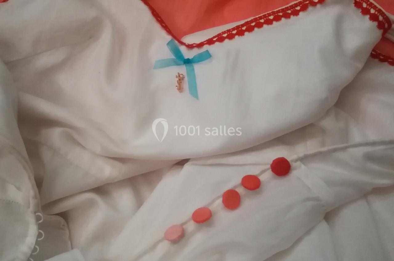 Robe blanche avec bordures rouges, ornée d'un nœud bleu et de boutons rouges, posée sur un tissu orange.