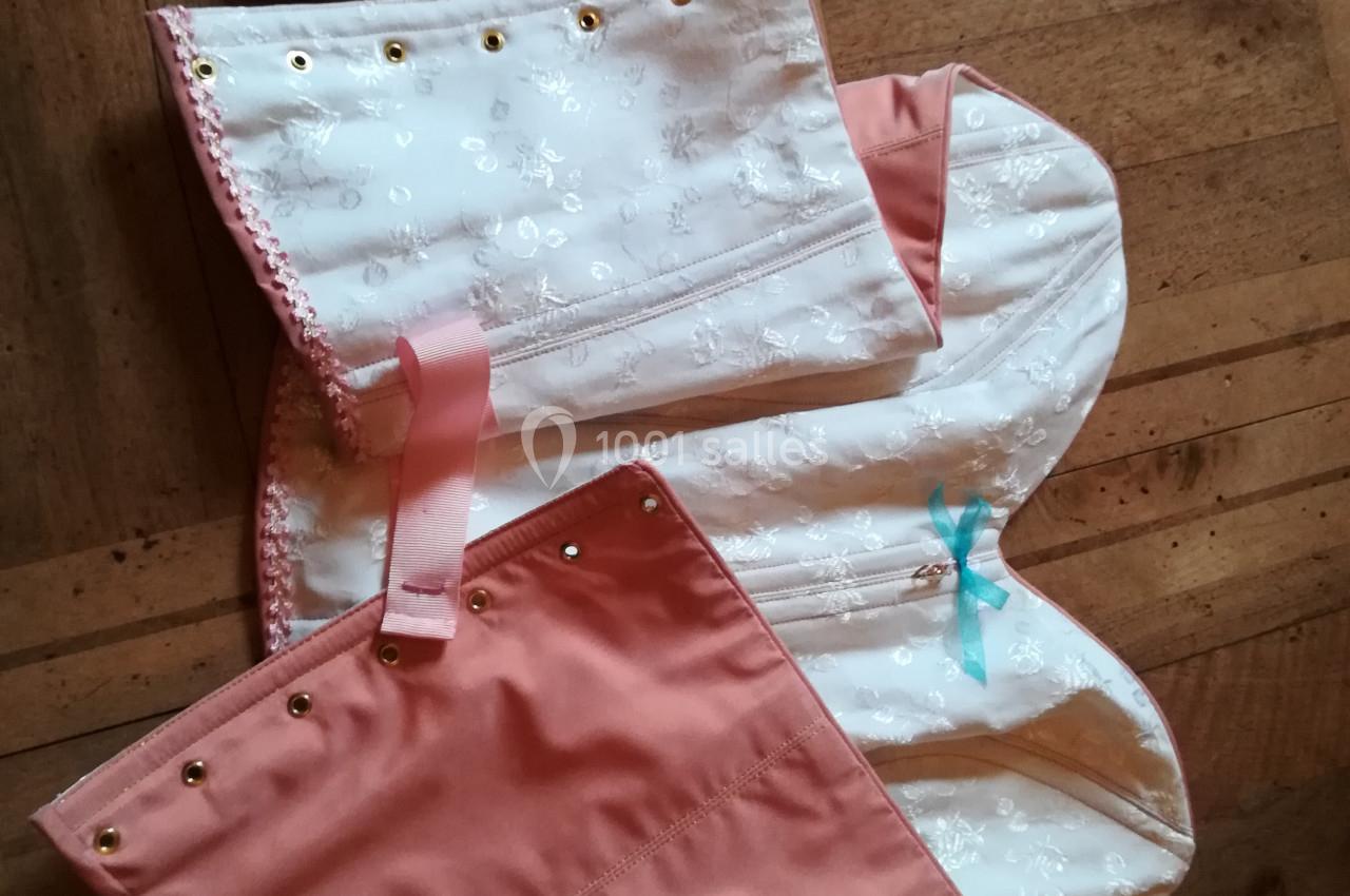 Corset en tissu rose et blanc à motifs floraux, avec lacets et œillets dorés, posé sur un sol en bois.