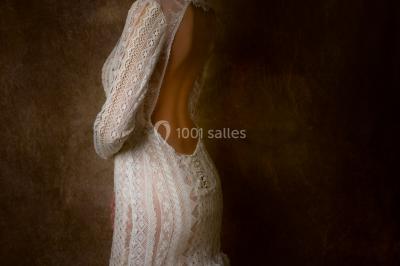 Mannequin vêtu d'une robe en dentelle blanche avec un voile noir couvrant la tête, devant une porte en bois clair.