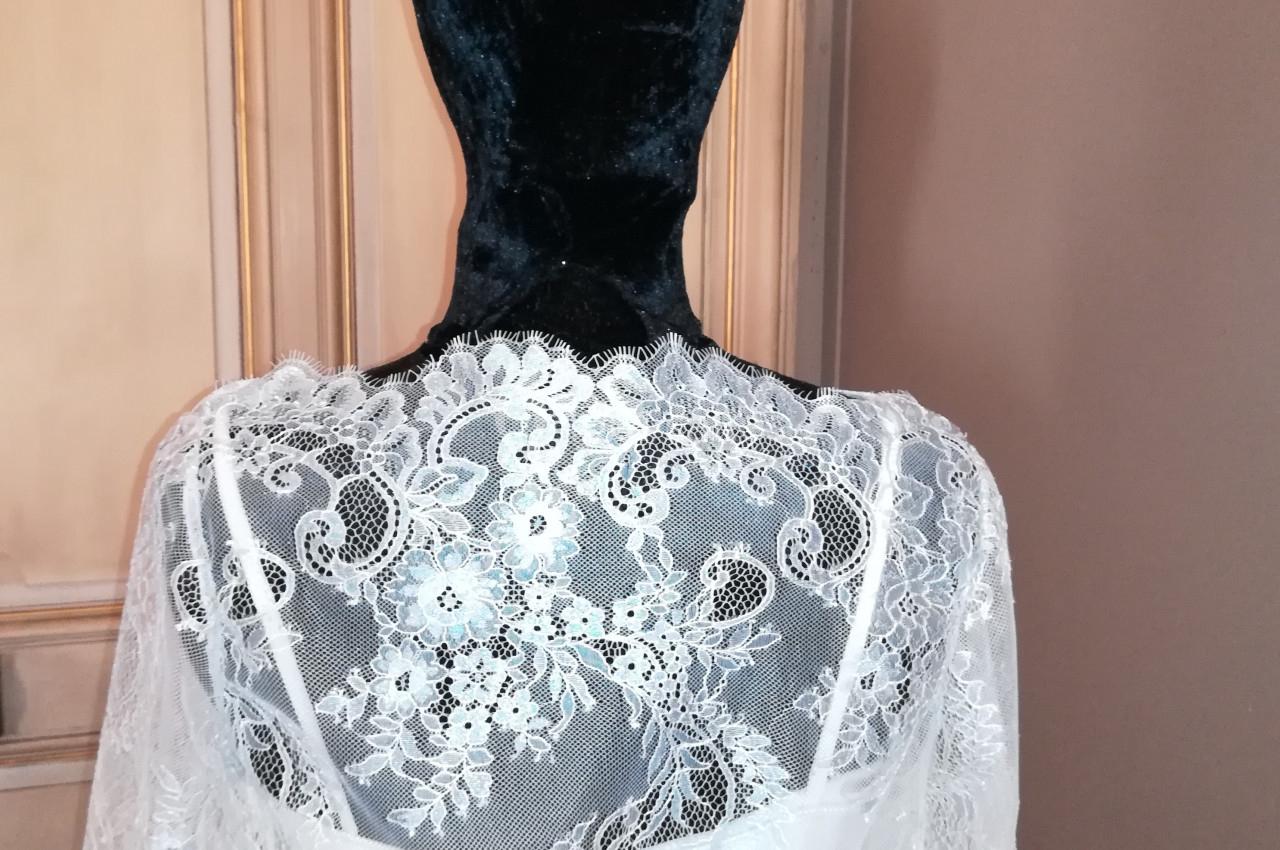 Mannequin vêtu d'une robe en dentelle blanche avec un voile noir couvrant la tête, devant une porte en bois clair.
