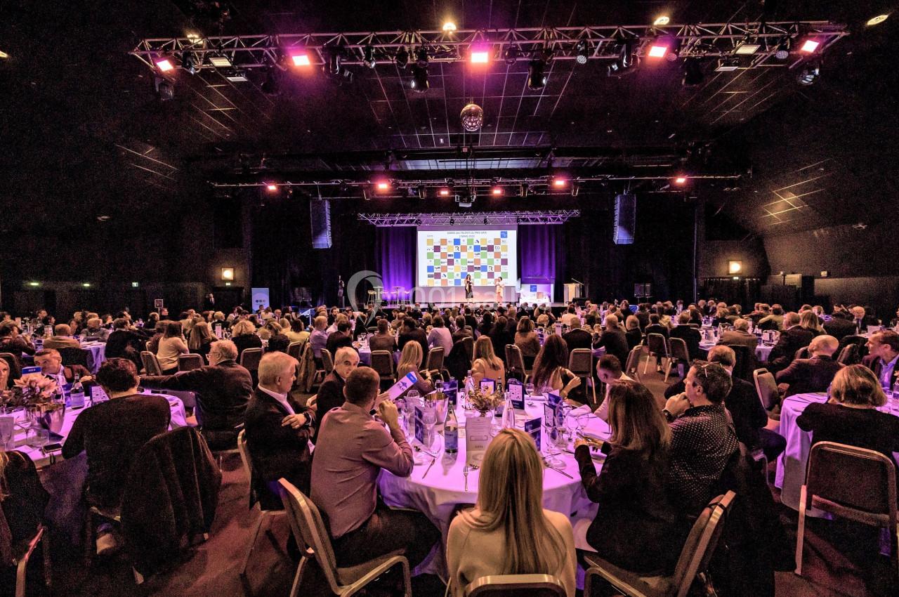 Salle de conférence remplie de participants assis à des tables rondes, face à une scène éclairée avec un écran coloré.