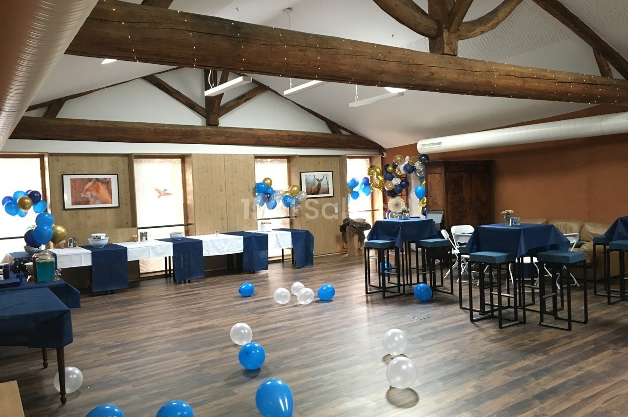 Salle décorée pour une fête avec ballons bleus et blancs, tables dressées et poutres apparentes au plafond.