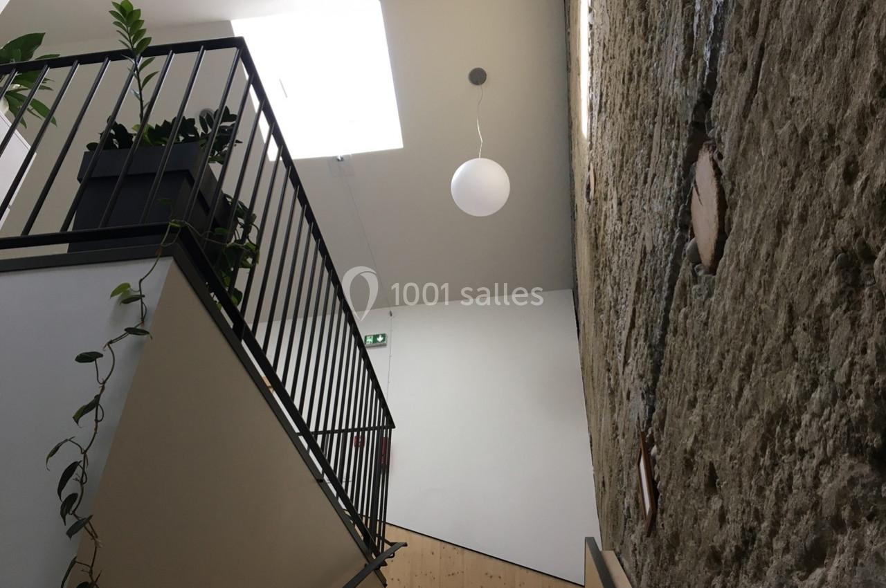 Escalier intérieur avec garde-corps en métal, mur en pierre apparente et éclairage naturel provenant d'un puits de lumière.