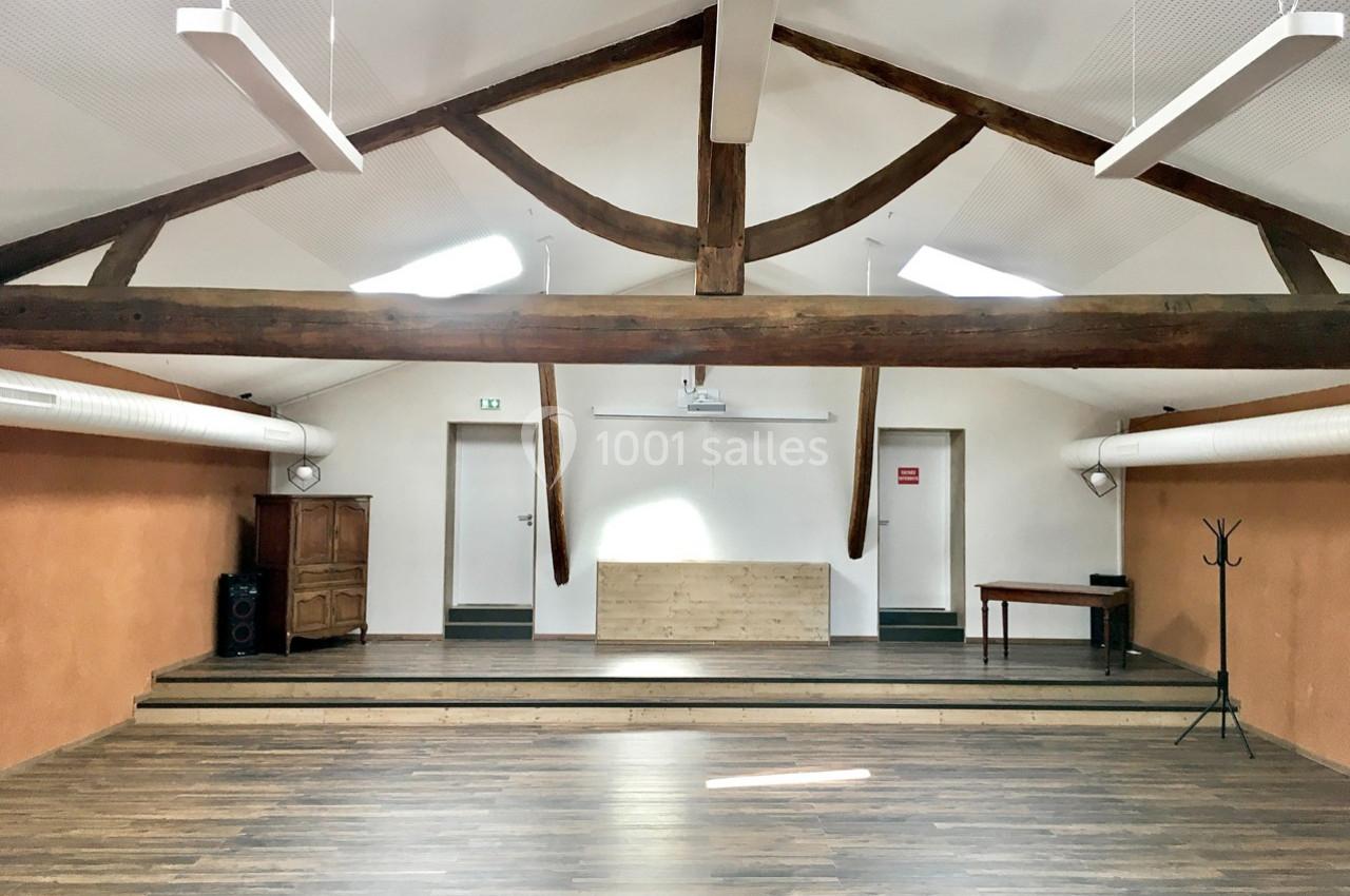 Salle lumineuse avec poutres apparentes, parquet en bois, estrade, mobilier minimaliste et éclairage suspendu.