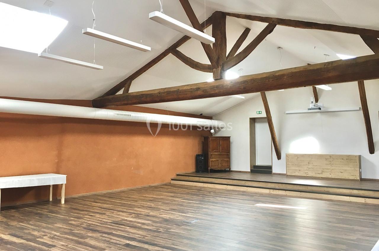 Salle lumineuse avec parquet en bois, poutres apparentes, murs orange et blancs, et une petite estrade en fond.