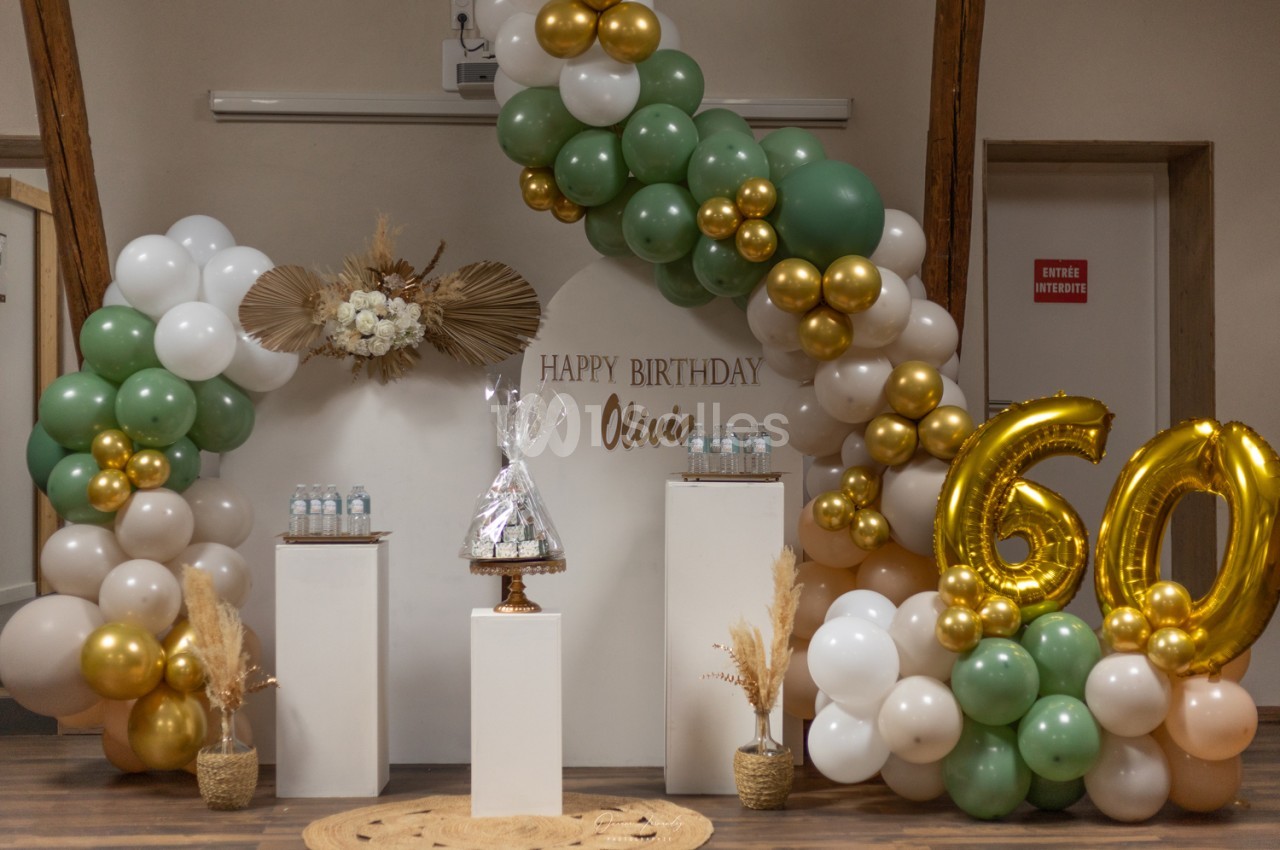 Décoration d'anniversaire avec arche de ballons verts, blancs, beige et dorés, chiffres dorés « 60 » et panneau « Happy Birthday » sur fond clair. Wait: must not transcribe text incrusted in image? The "Happy Birthday" banner is text on a backdrop; rule