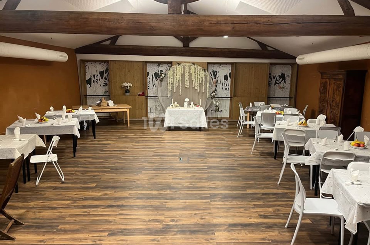 Grande salle aux poutres apparentes, dressée de tables rondes et carrées nappées de blanc, avec un espace décoré de fleurs blanches en fond. Let me verify length: roughly 130 characters? Count: "Grande salle aux poutres apparentes, dressée de tables