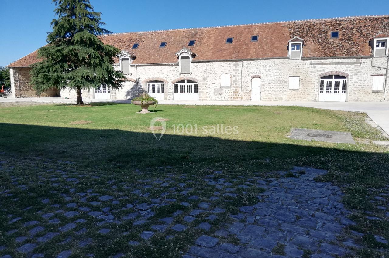 Bâtiment en pierre avec toit en tuiles rouges, grandes fenêtres et pelouse arborée au premier plan.