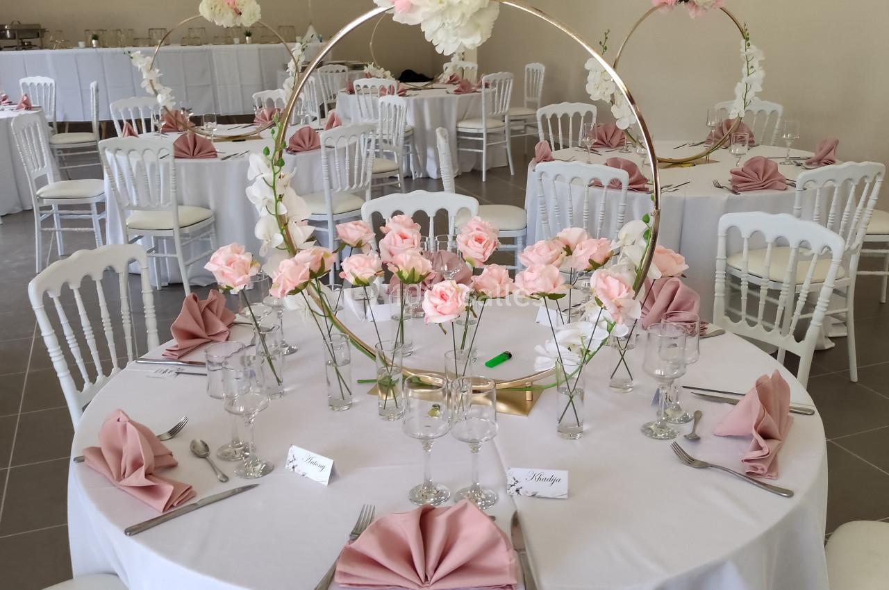 Salle de réception décorée avec des tables rondes, nappes blanches, serviettes roses et centres de table fleuris.