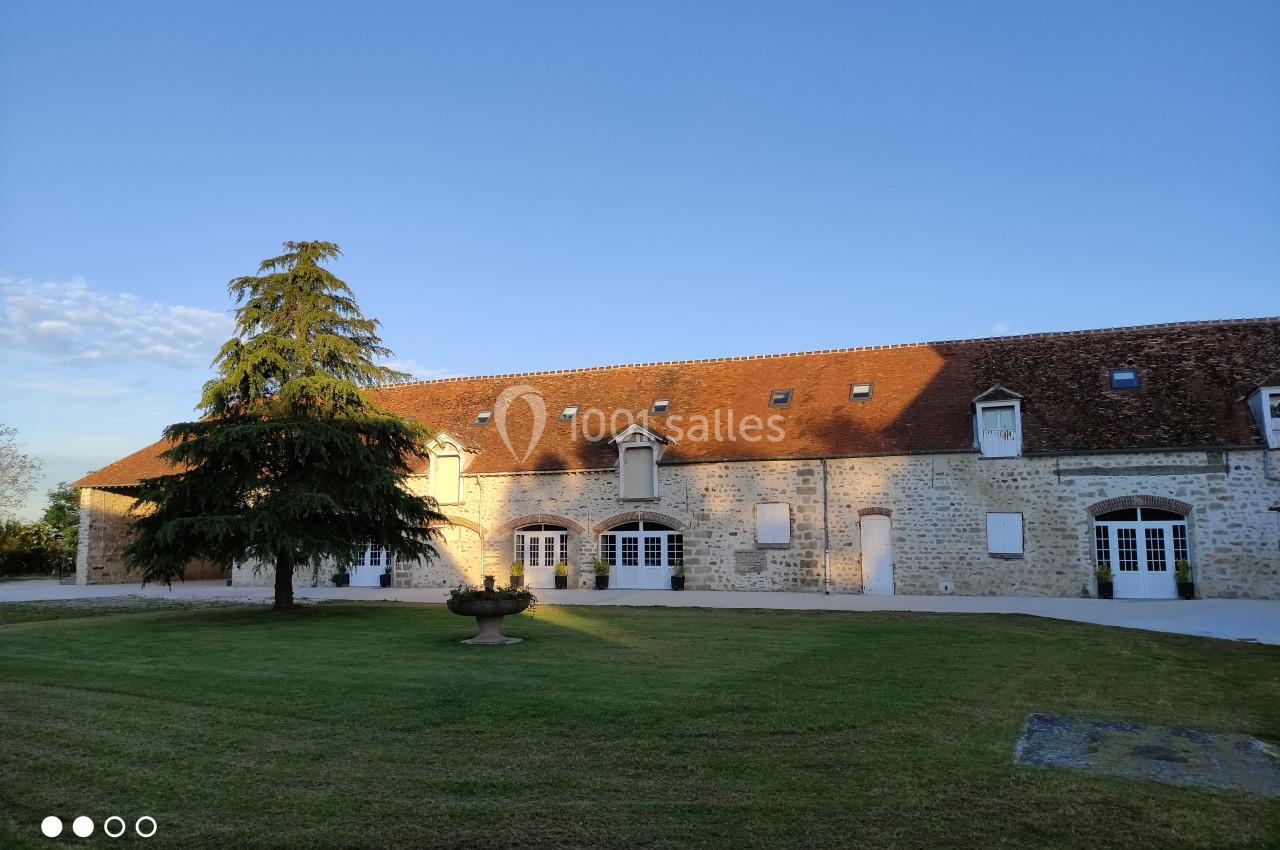 Bâtiment en pierre avec toit en tuiles, grandes fenêtres blanches et pelouse arborée au premier plan.