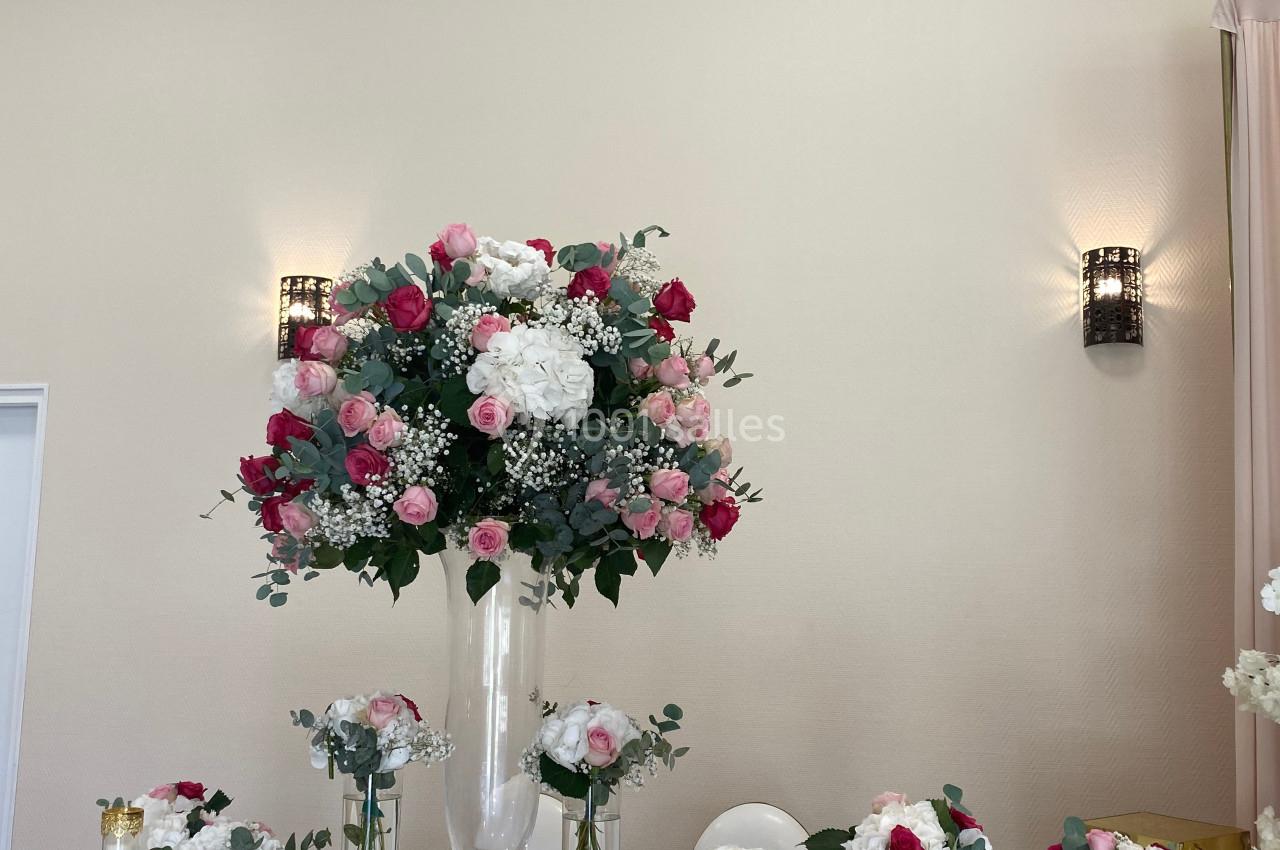Arrangement floral avec roses roses et blanches dans un vase central, entouré de petits bouquets sur une table blanche.