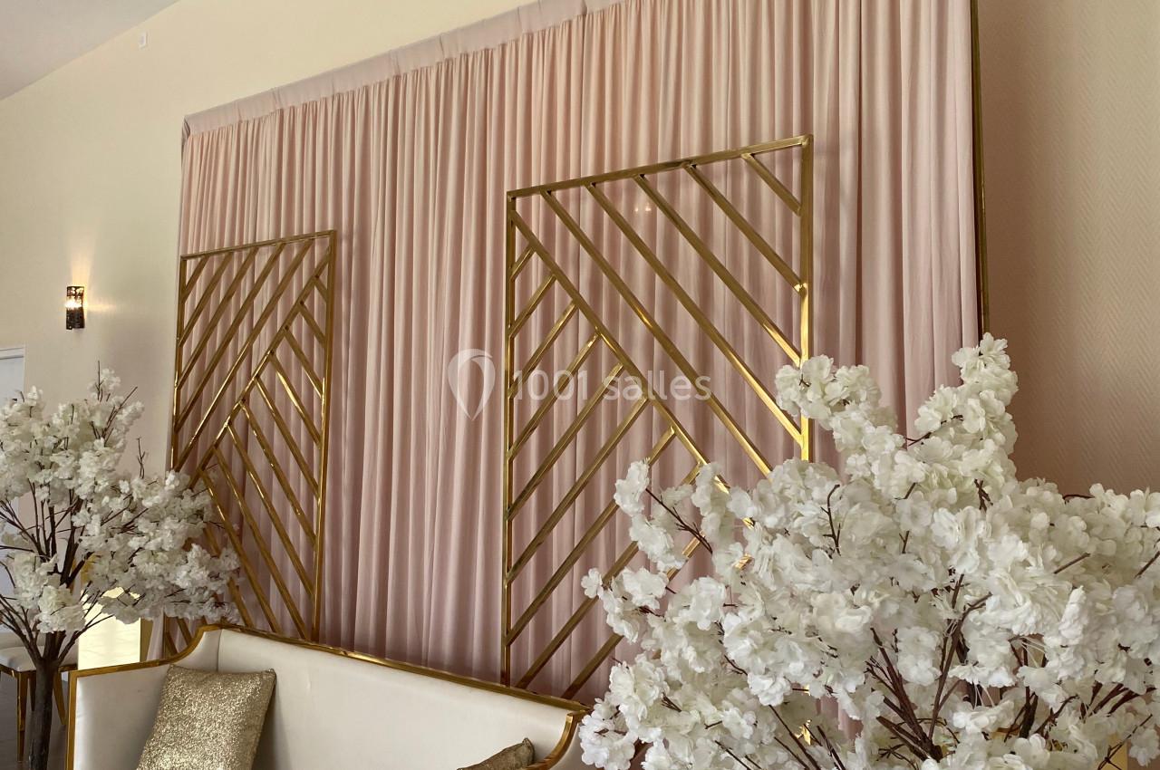 Mur décoratif avec rideaux roses, structures dorées, canapé blanc, coussins dorés et arbres fleuris blancs.