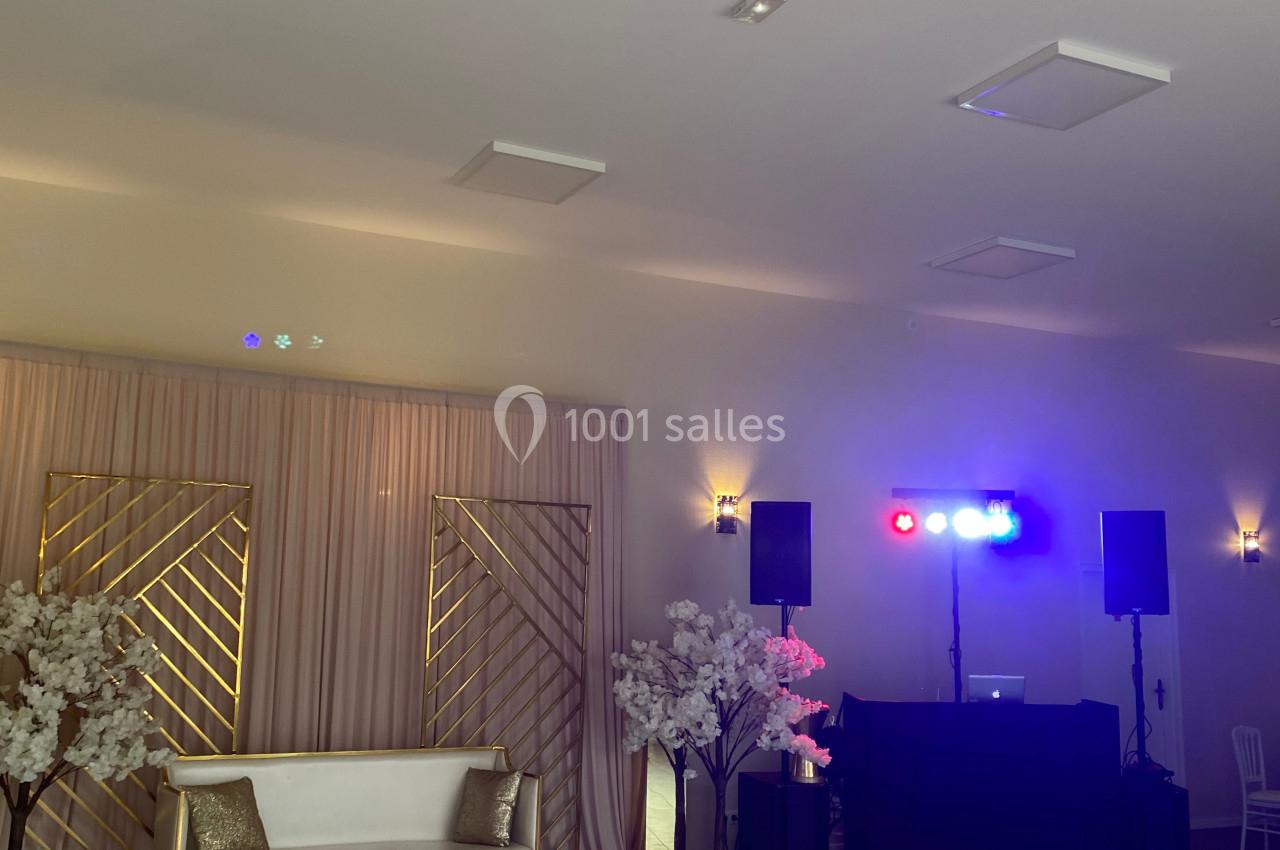 Salle de réception avec canapé, décor floral, rideaux beiges, éclairage coloré et espace DJ équipé.