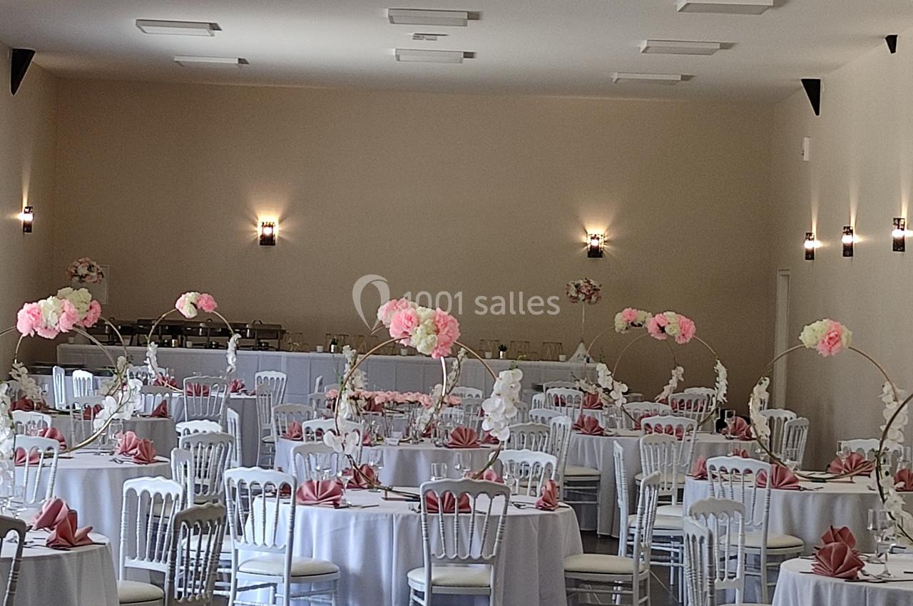 Salle de réception décorée avec des tables rondes, nappes blanches, serviettes roses et centres de table fleuris.