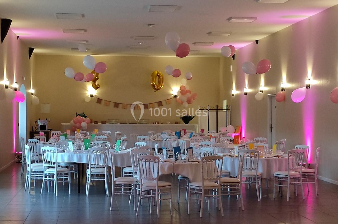 Salle décorée pour une fête avec des tables rondes, nappes blanches, chaises en bois et ballons roses et blancs suspendus.