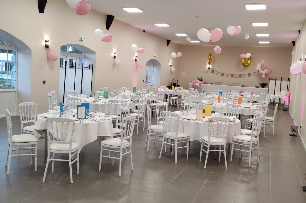 Salle décorée pour une fête, avec des tables rondes dressées, des chaises blanches et des ballons roses et blancs suspendus.