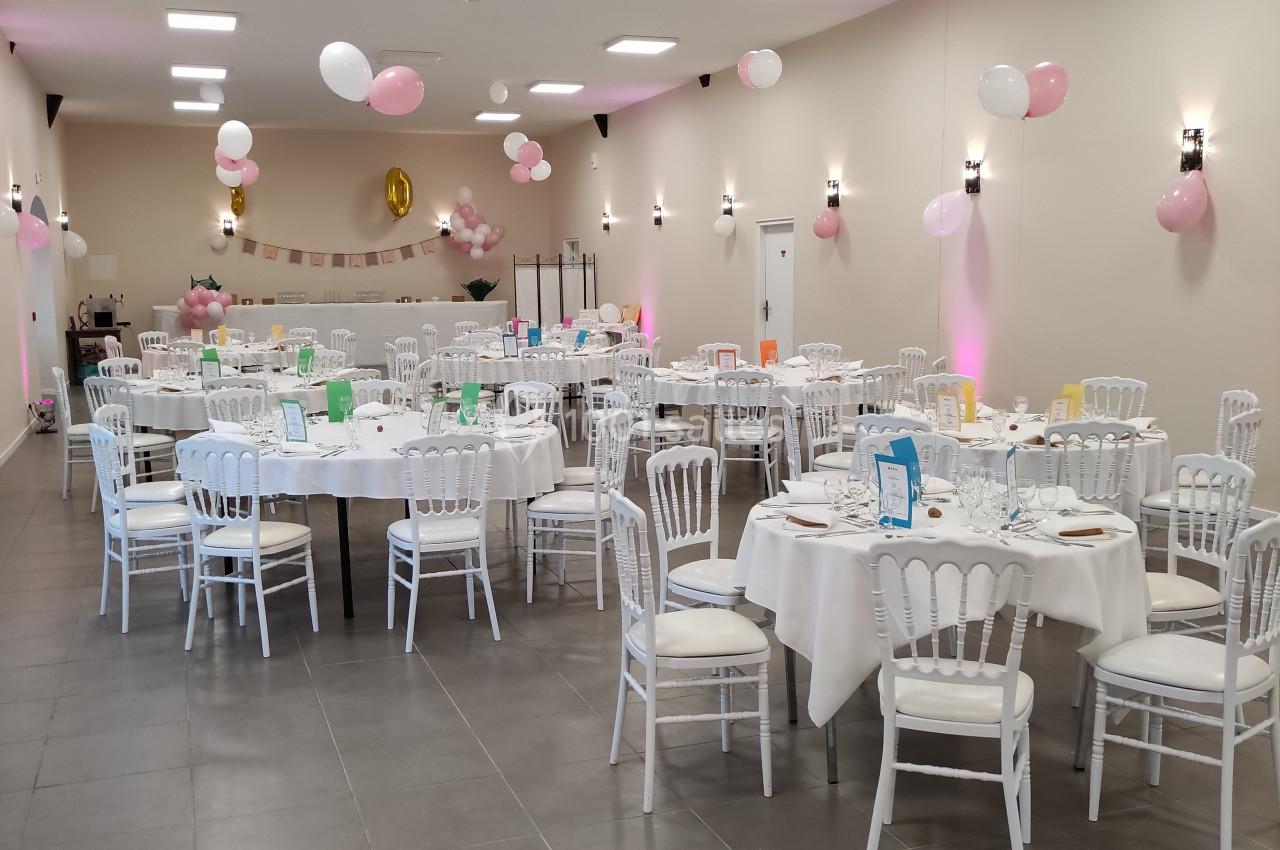 Salle décorée pour un événement avec des tables rondes dressées, des chaises blanches et des ballons roses et blancs.